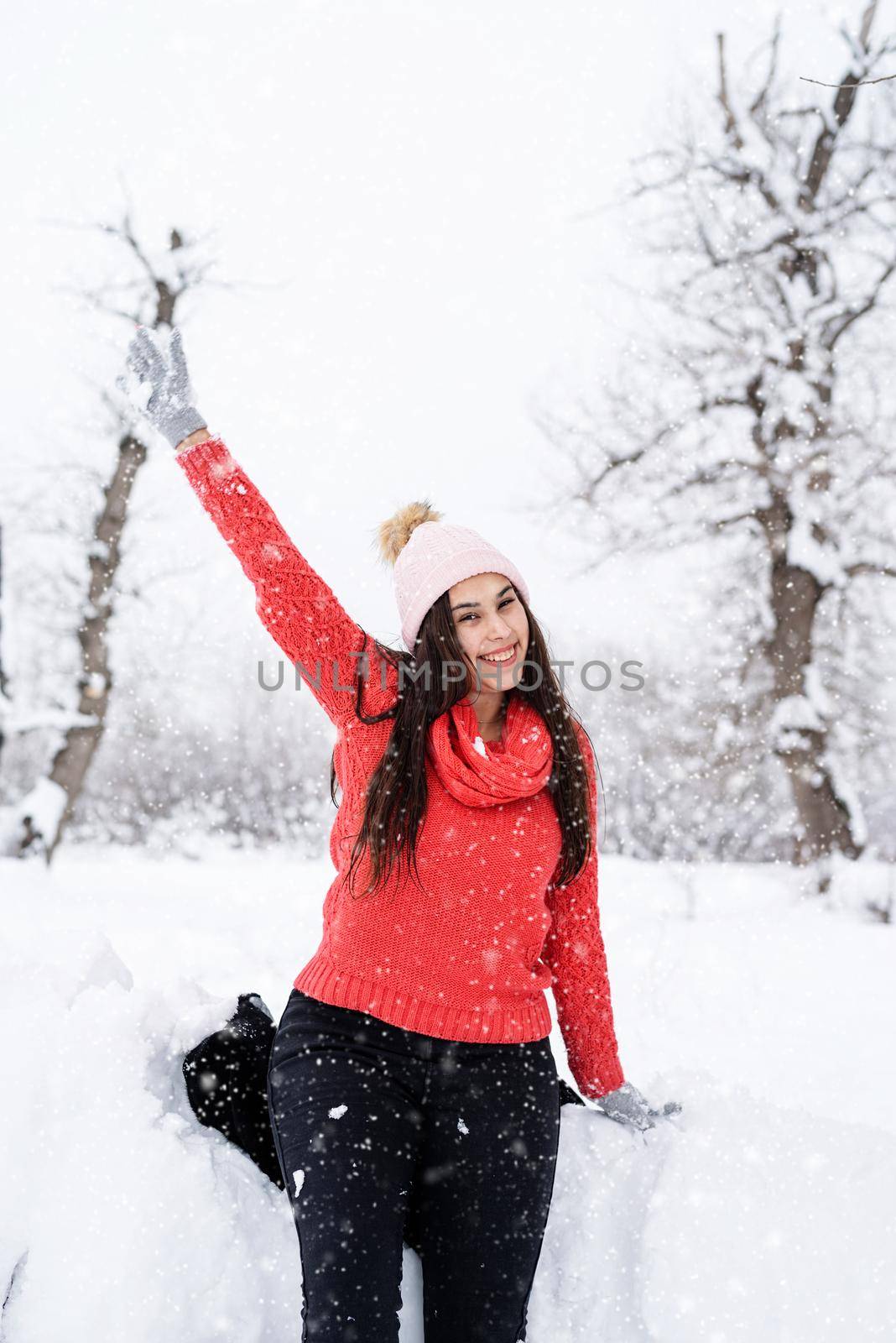 Winter season. Portrait of a beautiful smiling woman in red sweater and hat in snowy park waving hello
