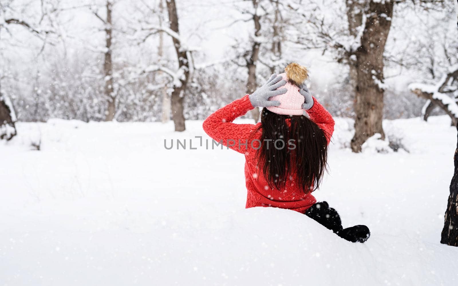 Winter season. Rear view of brunette woman sitting in snowy park in snowfall putting on her hat