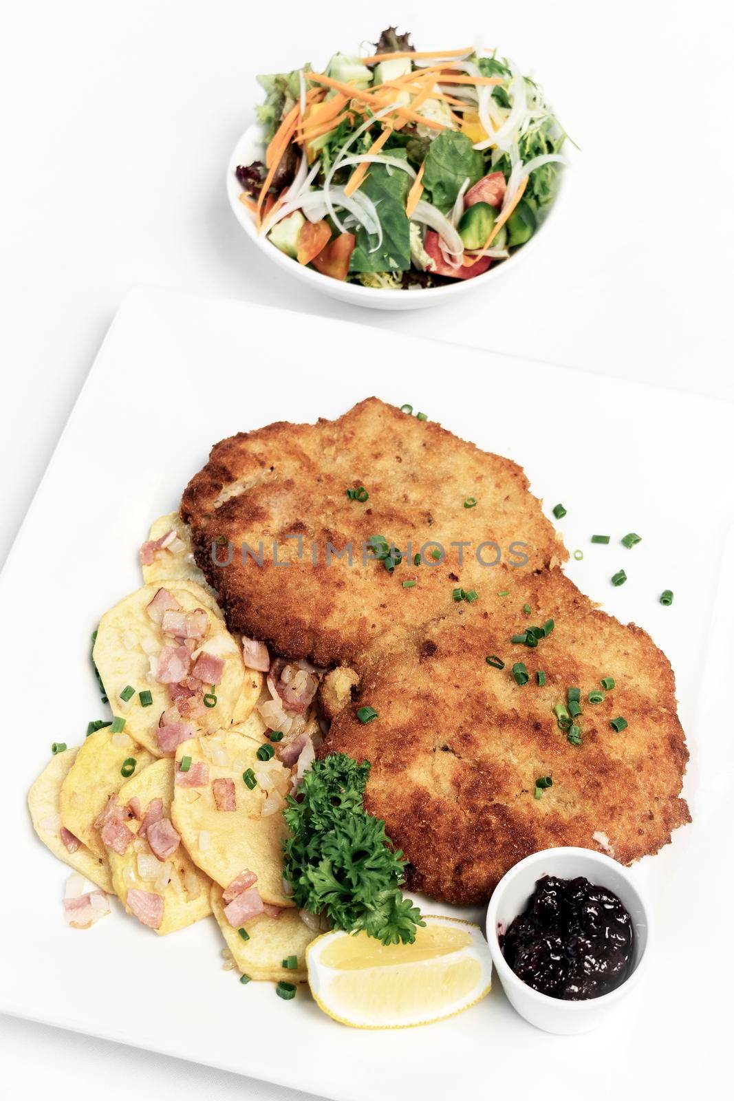 german breaded organic pork schnitzel with bacon fried potatoes cranberry sauce and salad on white studio background