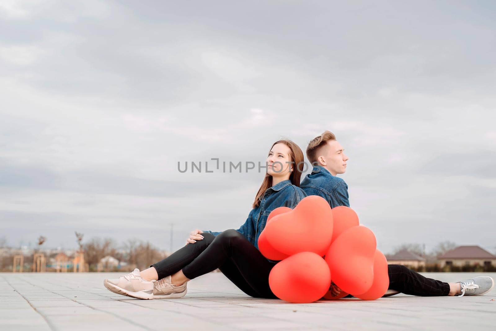 Young loving smiling couple sitting back to back in the street holding a pile of red balloons spending time together