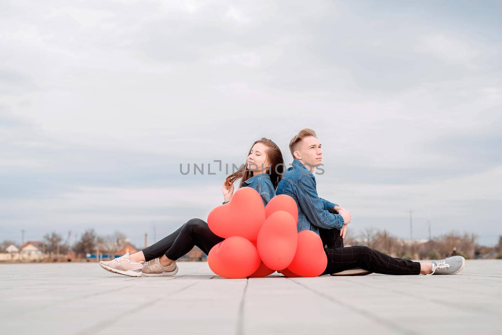 Young loving couple sitting back to back in the street holding a pile of red balloons spending time together by Desperada