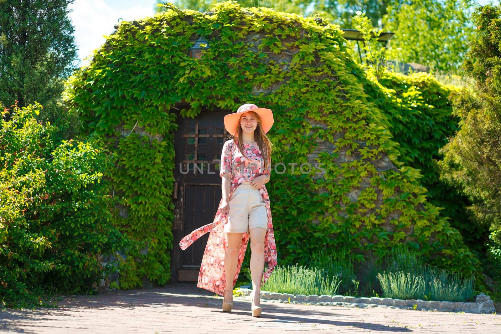 A charming girl in a light summer sundress and a pareo hat is walking in a green park. Enjoys warm sunny summer days.