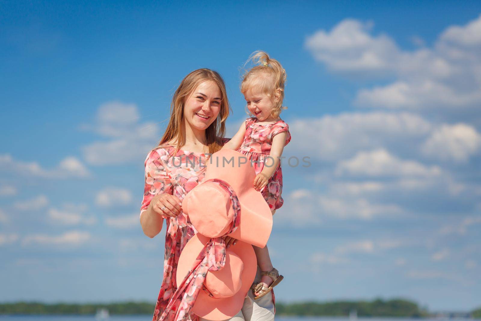 A charming girl in a light summer sundress walks on the sandy beach with her little daughter. Enjoys warm sunny summer days.
