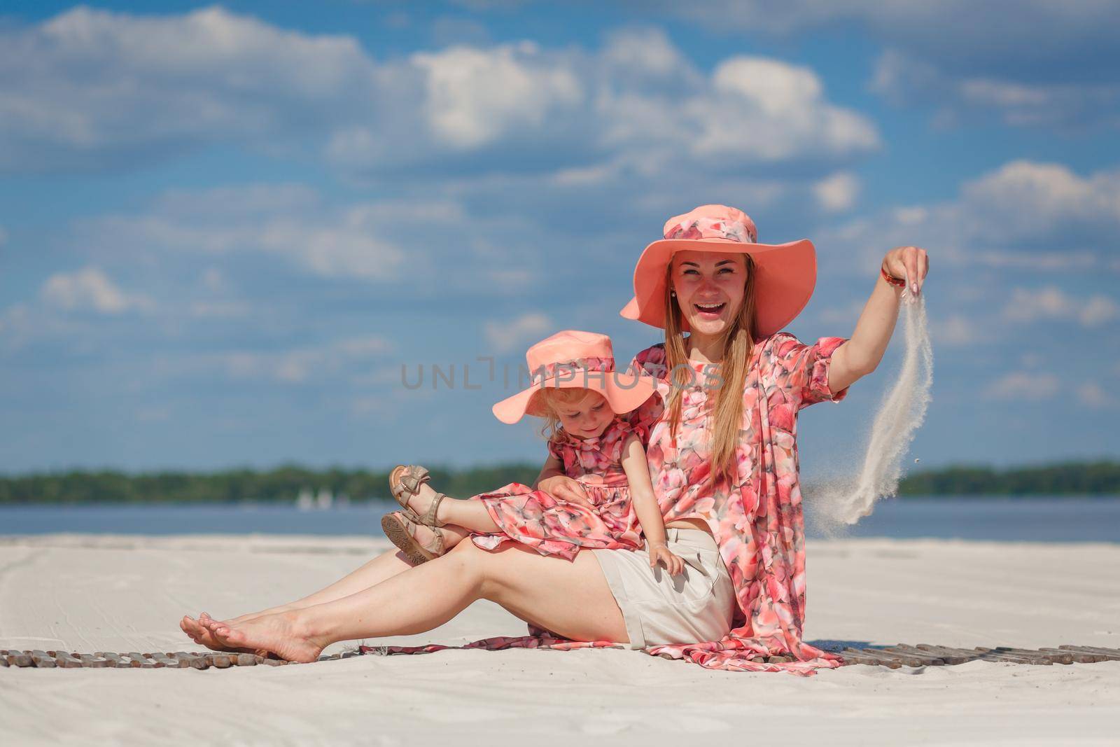 A little girl with her mother in matching beautiful sundresses plays in the sand on the beach. Stylish family look. by Try_my_best