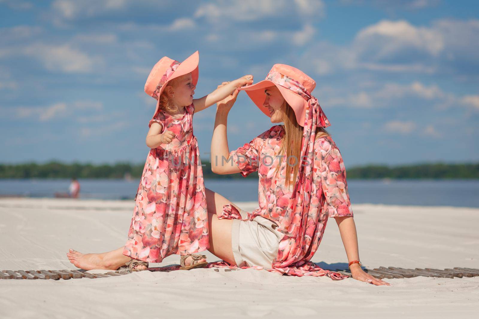 A little girl with her mother in matching beautiful sundresses plays in the sand on the beach. Stylish family look. by Try_my_best