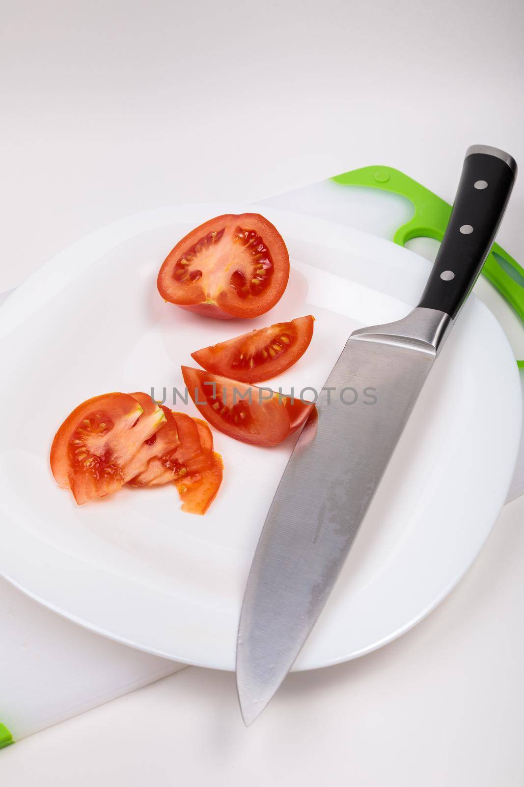 On a cutting board is a fresh red cut tomato and a large chef's knife. On an isolated white background.