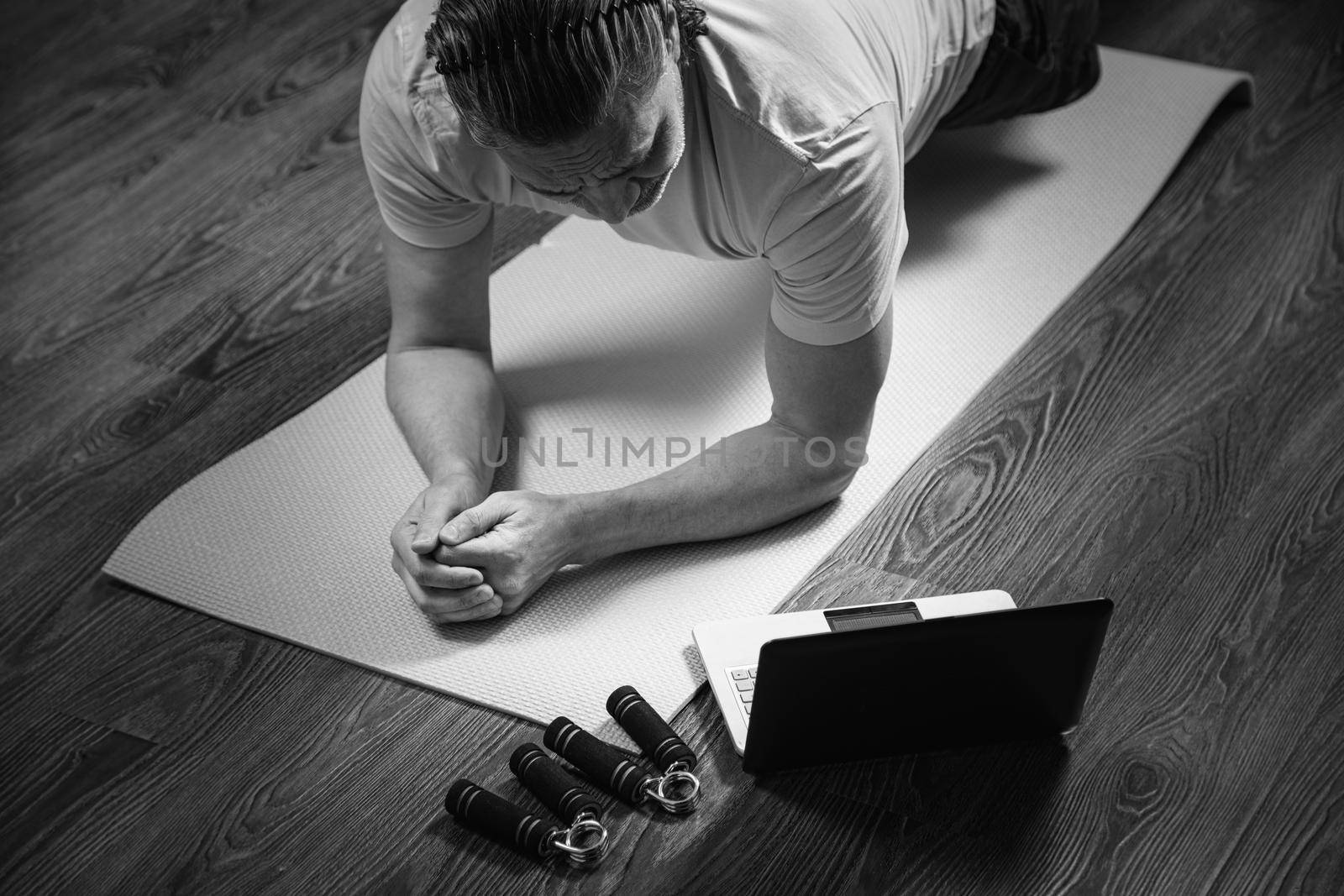 50-year-old man performs exercises while lying on the rug at home, looking at the computer. During a pandemic, a person trains in an apartment via the Internet.