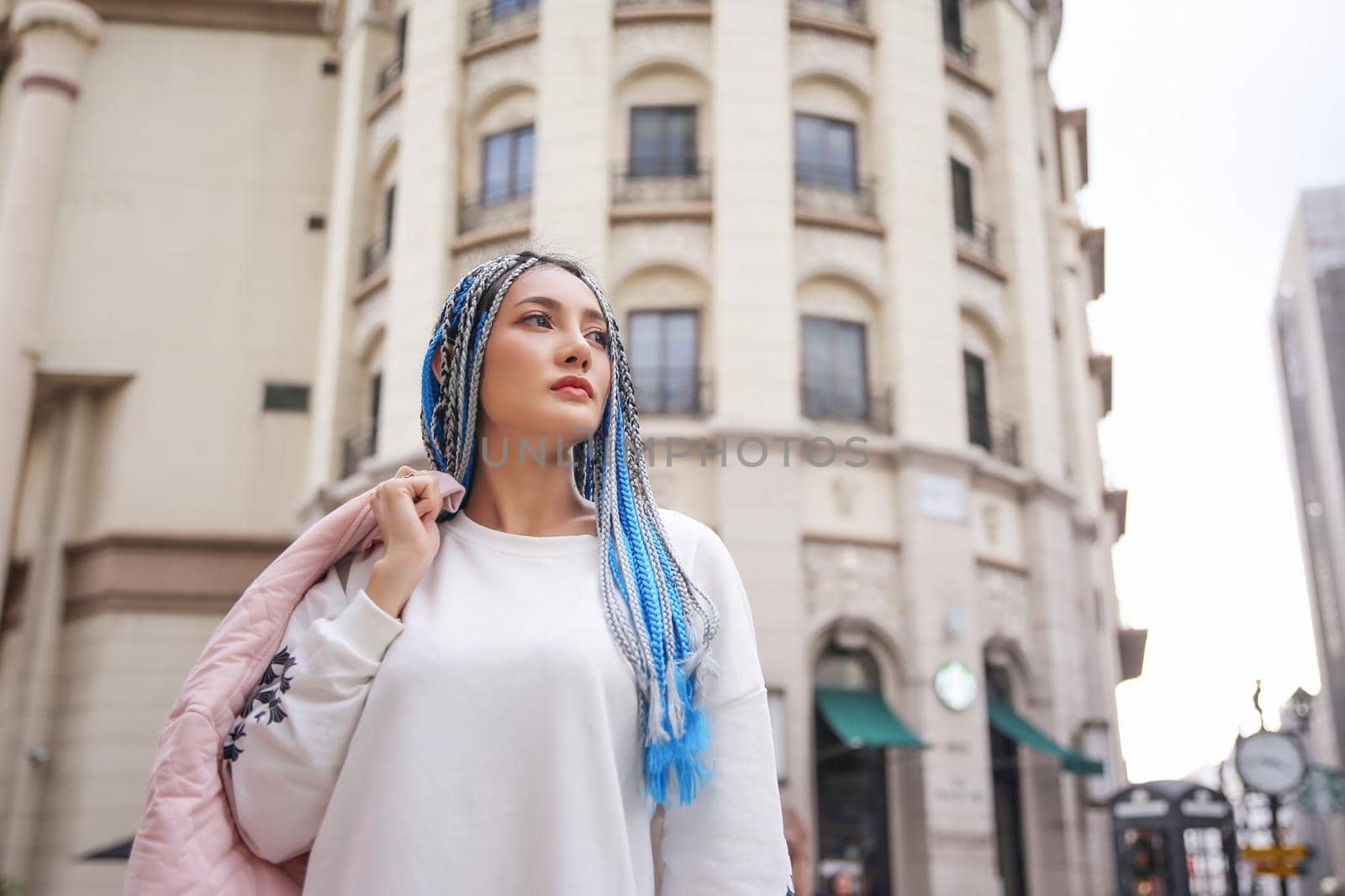 Happy stylish young hipster woman with long blue hair pink jacket, hat on the street in urban city.