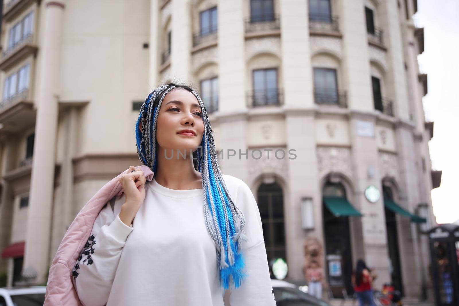 Happy stylish young hipster woman with long blue hair pink jacket, hat on the street in urban city.