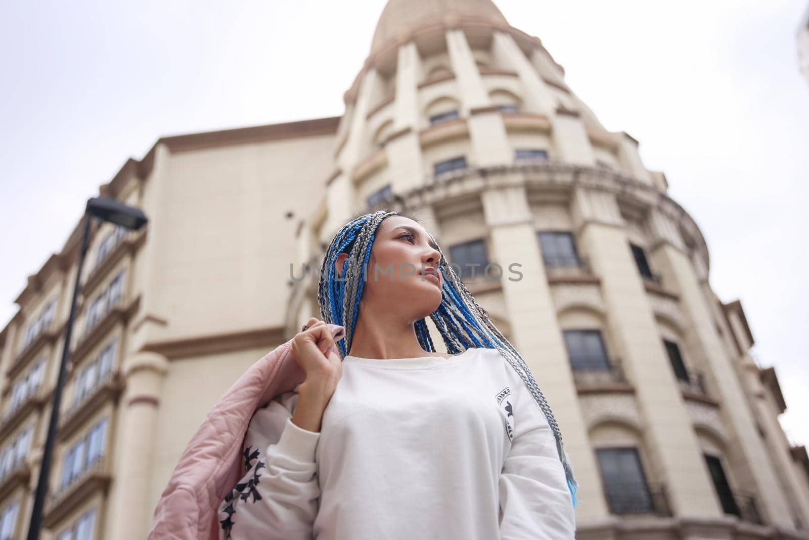 Happy stylish young hipster woman with long blue hair pink jacket, hat on the street in urban city.