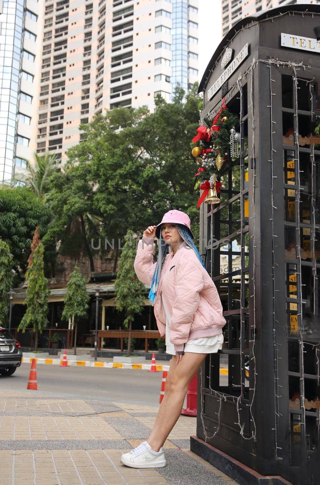 Happy stylish young hipster woman with long blue hair pink jacket, hat on the street in urban city.