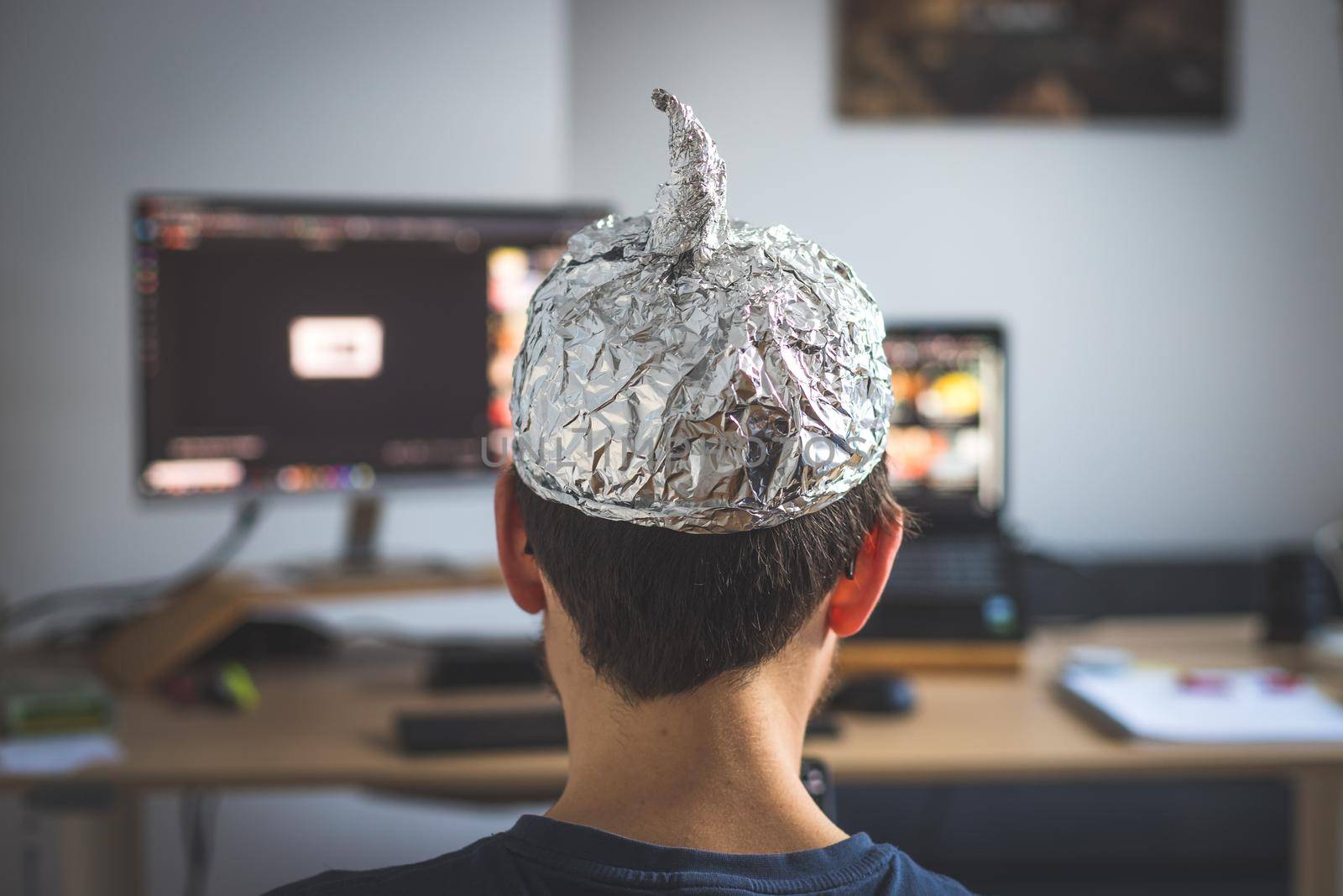 Conspiracy theory concept: young man is wearing aluminum head, sitting in front of the pc watching videos by Daxenbichler