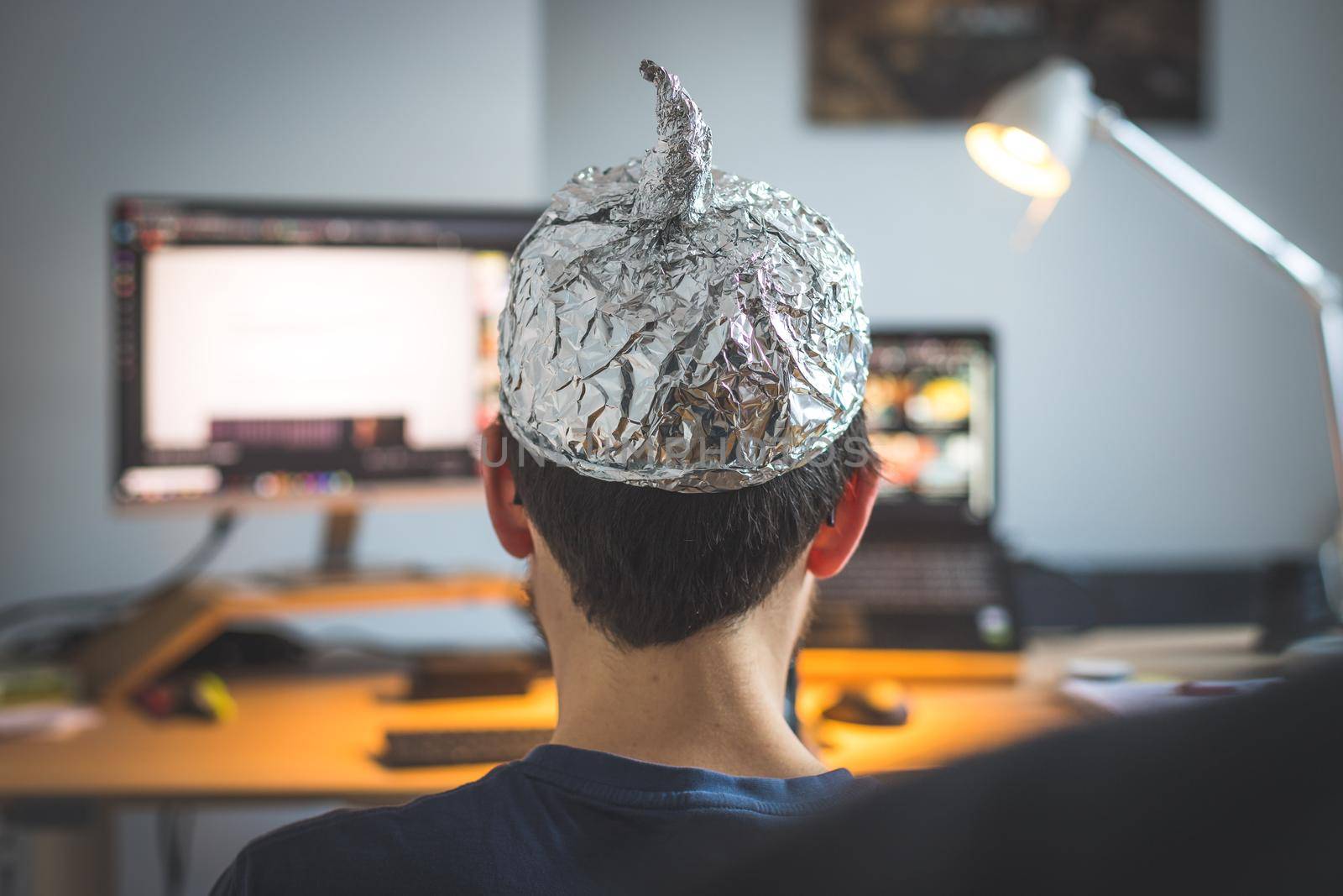 Conspiracy theory concept: young man is wearing aluminum head, sitting in front of the pc watching videos by Daxenbichler