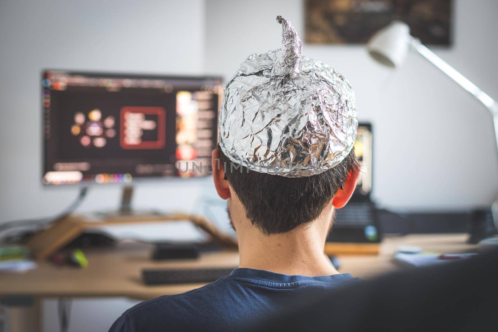 Conspiracy theory concept: young man is wearing aluminum head, sitting in front of the pc watching videos by Daxenbichler