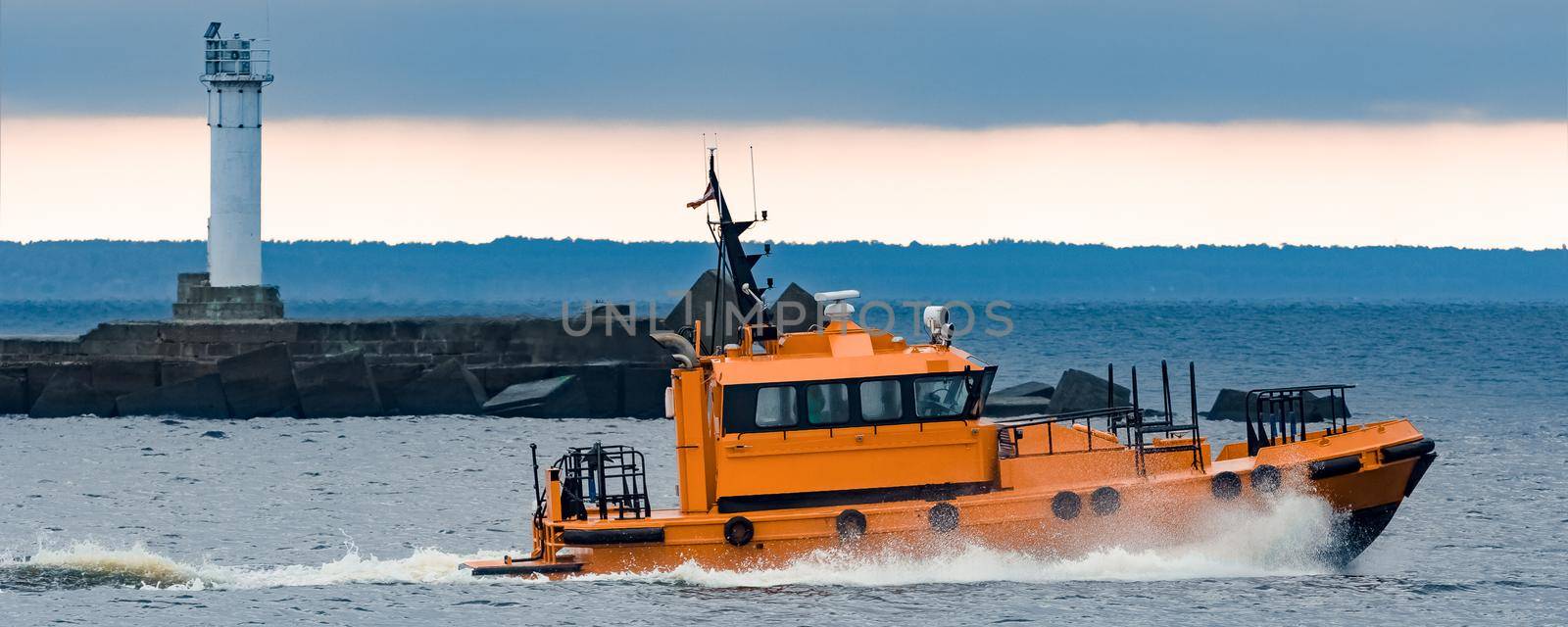 Orange pilot ship moving at speed past the lighthouse in Riga