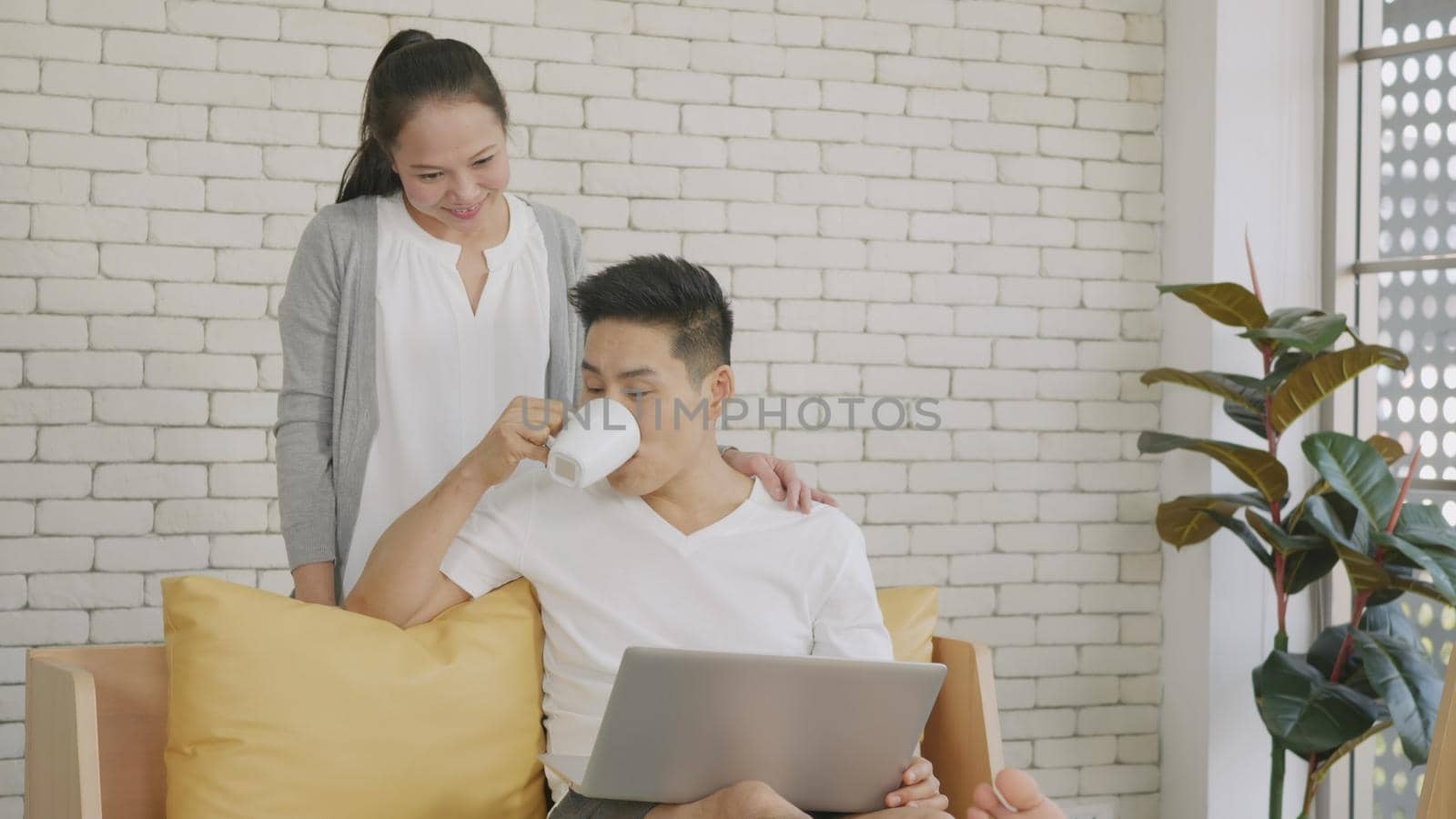 Happy Asian beautiful family couple husband and wife laughing sitting on sofa in the living room working with laptop computer at home. Woman brings coffee to the man during work