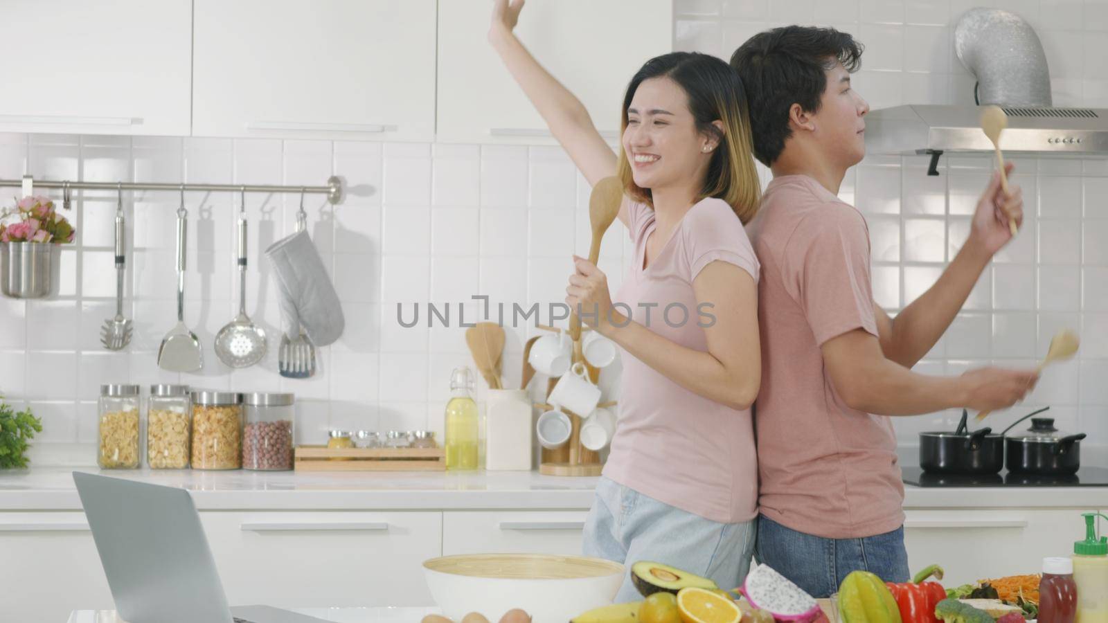 Happy Asian beautiful family couple husband and wife singing in kitchenware microphones in kitchen together having fun dance listen music at home. Two people dancing