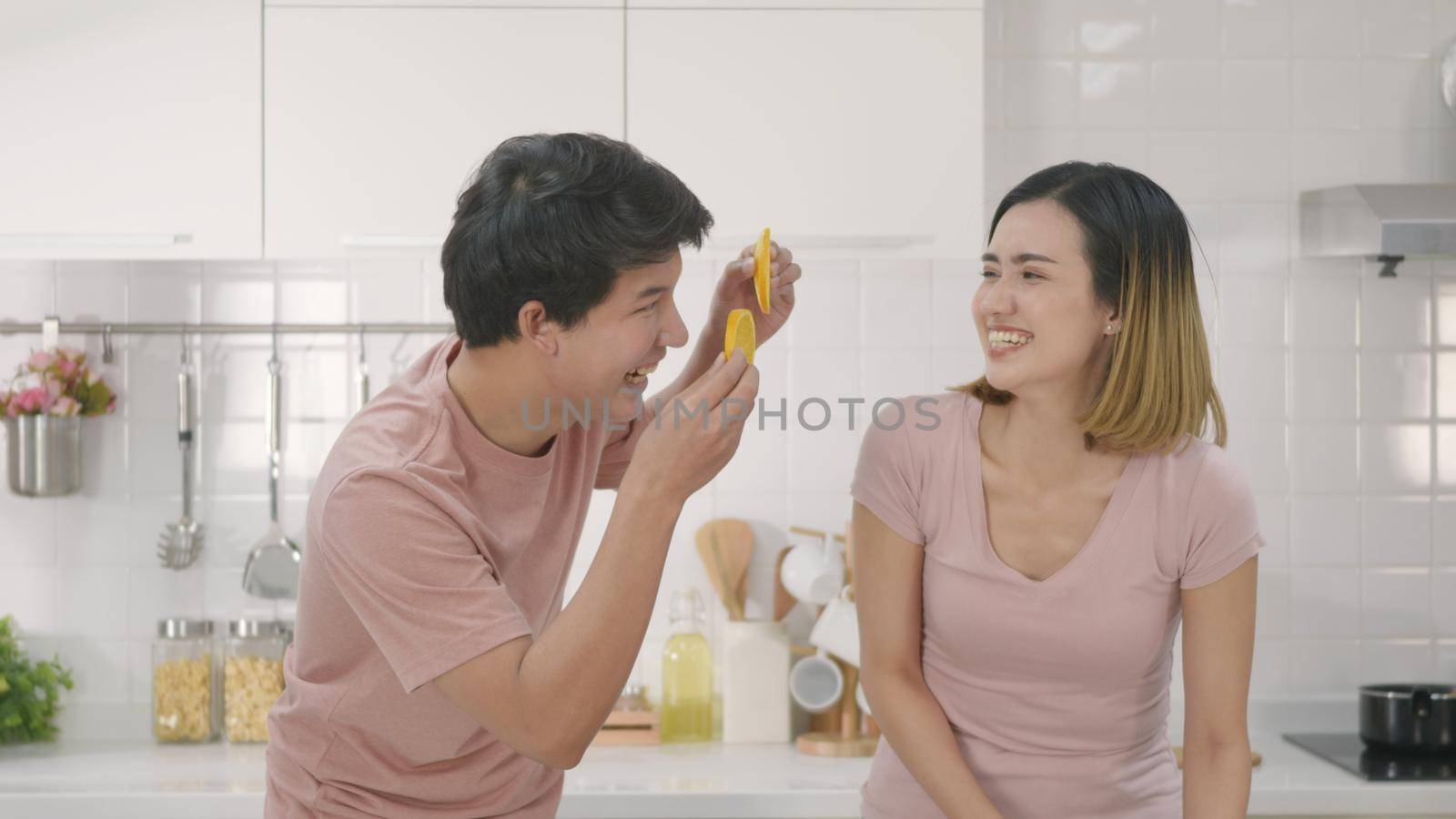 Happy Asian beautiful young family couple husband and wife enjoying smile and laugh holds a cut orange in front of the guy's eyes spending time together in kitchen at home.