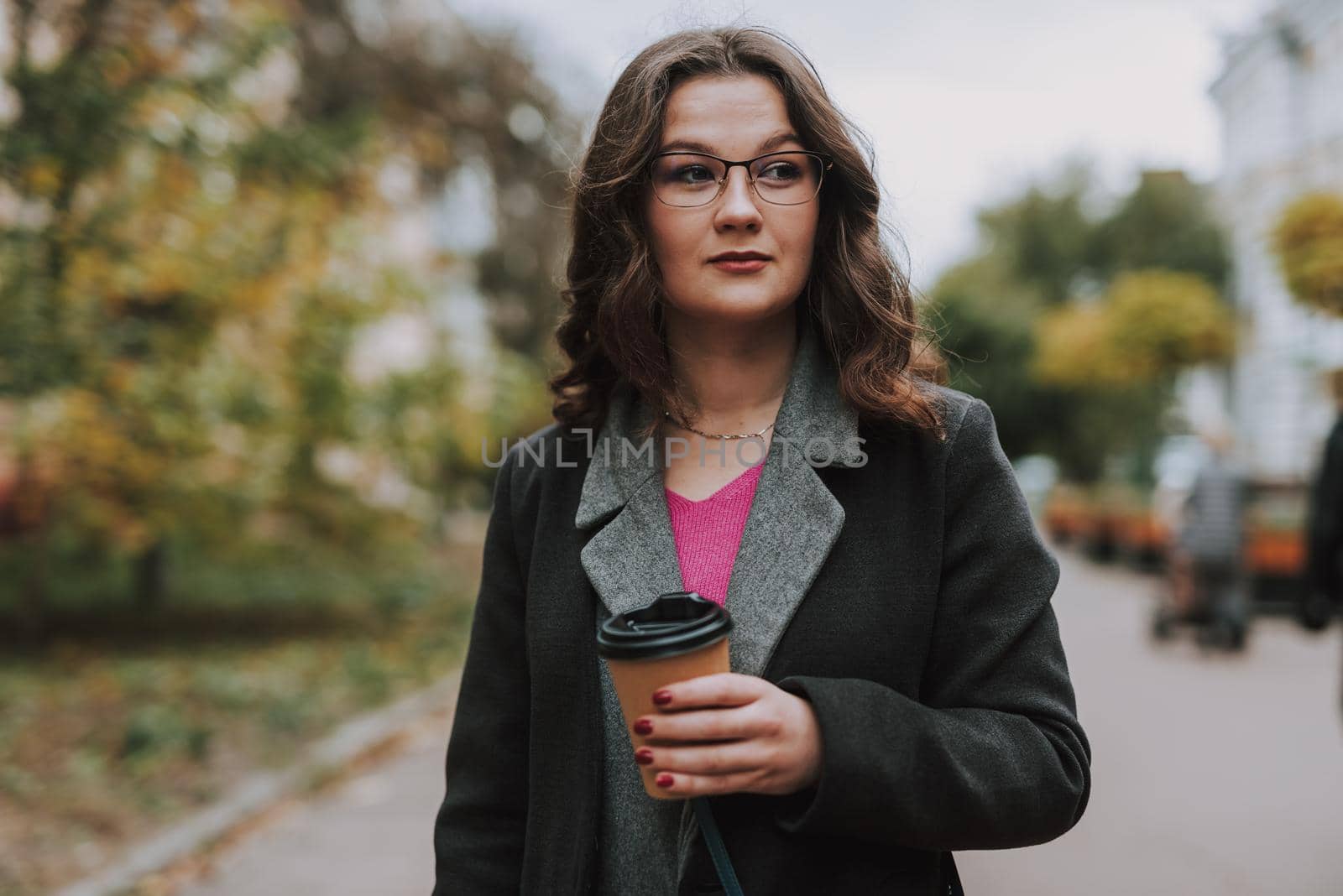 Calm beautiful young woman on autumn day drinking coffee in the street and looking into the distance