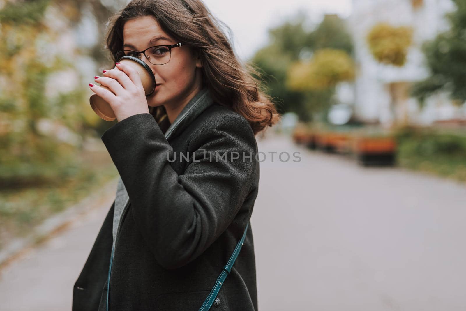 Attractive young lady in the empty street standing alone and looking calm while drinking taking a sip of coffee from a paper cup