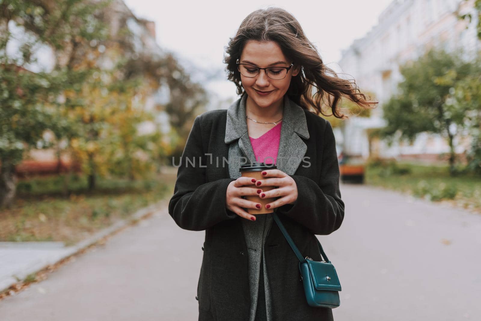 Pretty curly young woman smiling and looking at a paper cup of coffee in her hands while standing in the street