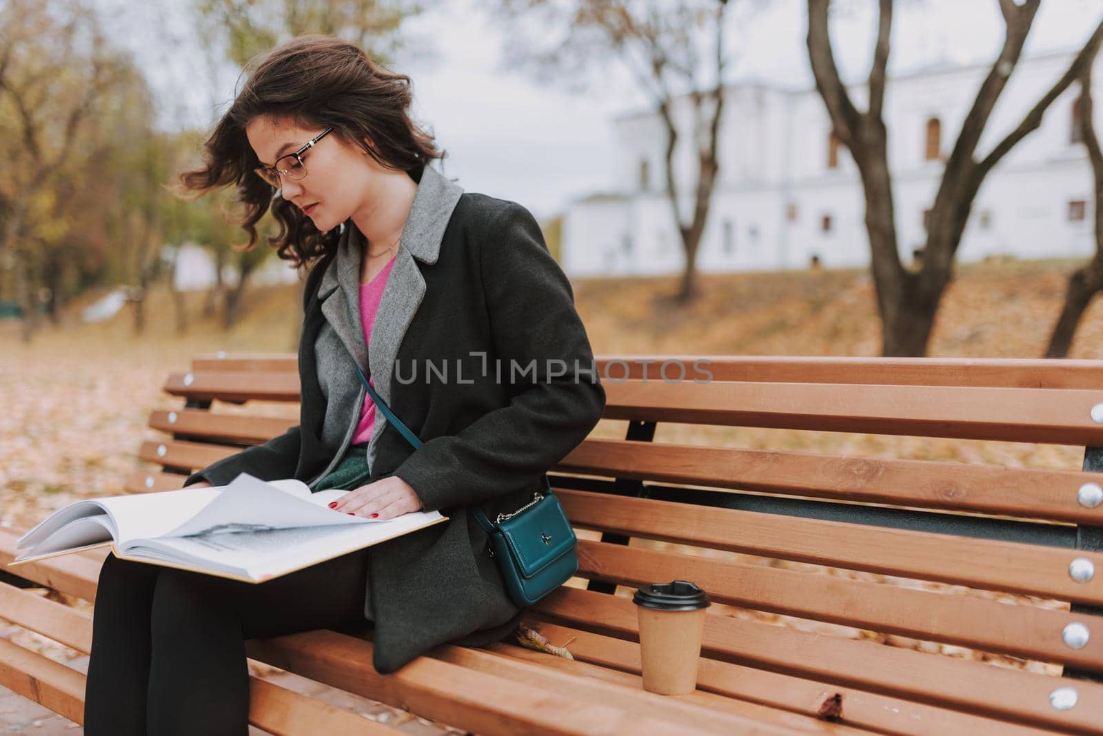 Beautiful female student wearing glasses and leafing through pages of book in a park while studying