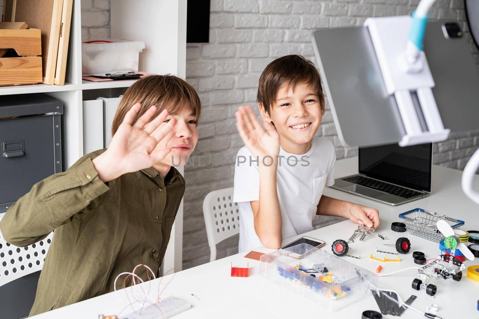 young boys having fun making robot cars having online class on digital tablet greeting teacher