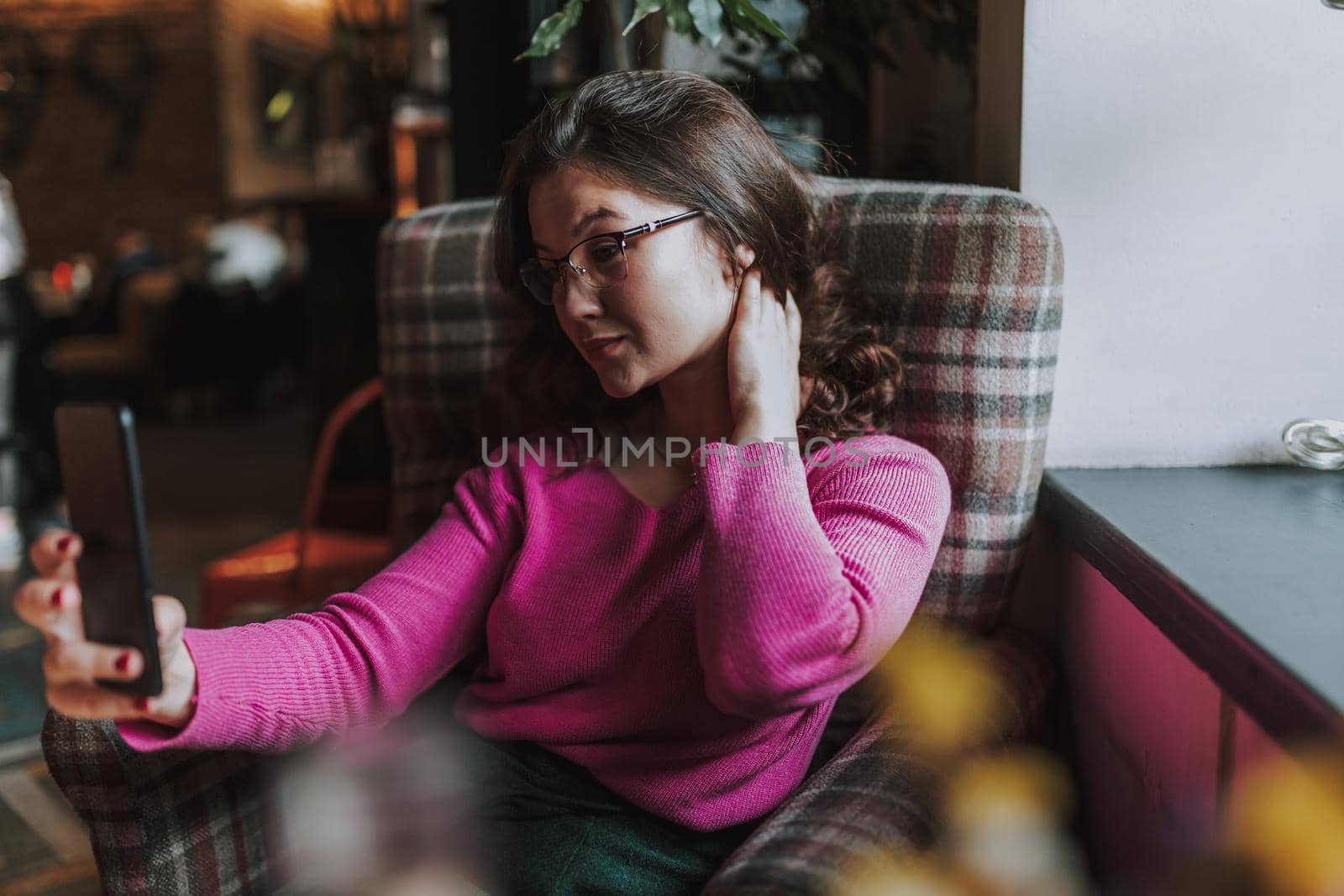 Smiling pretty young woman holding smartphone and taking selfie while resting in coffee house