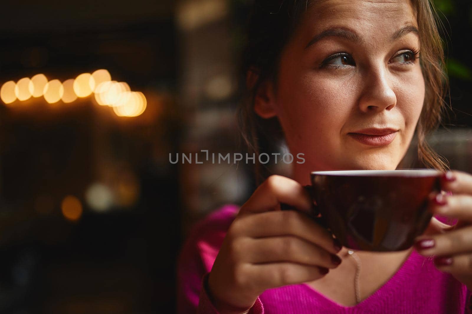 Thoughtful young woman sitting in coffee house and looking out the window while drinking tea