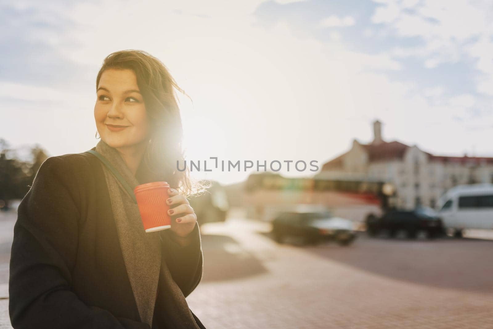 Waist up of happy pretty lady standing on the street while holding hot drink and looking away