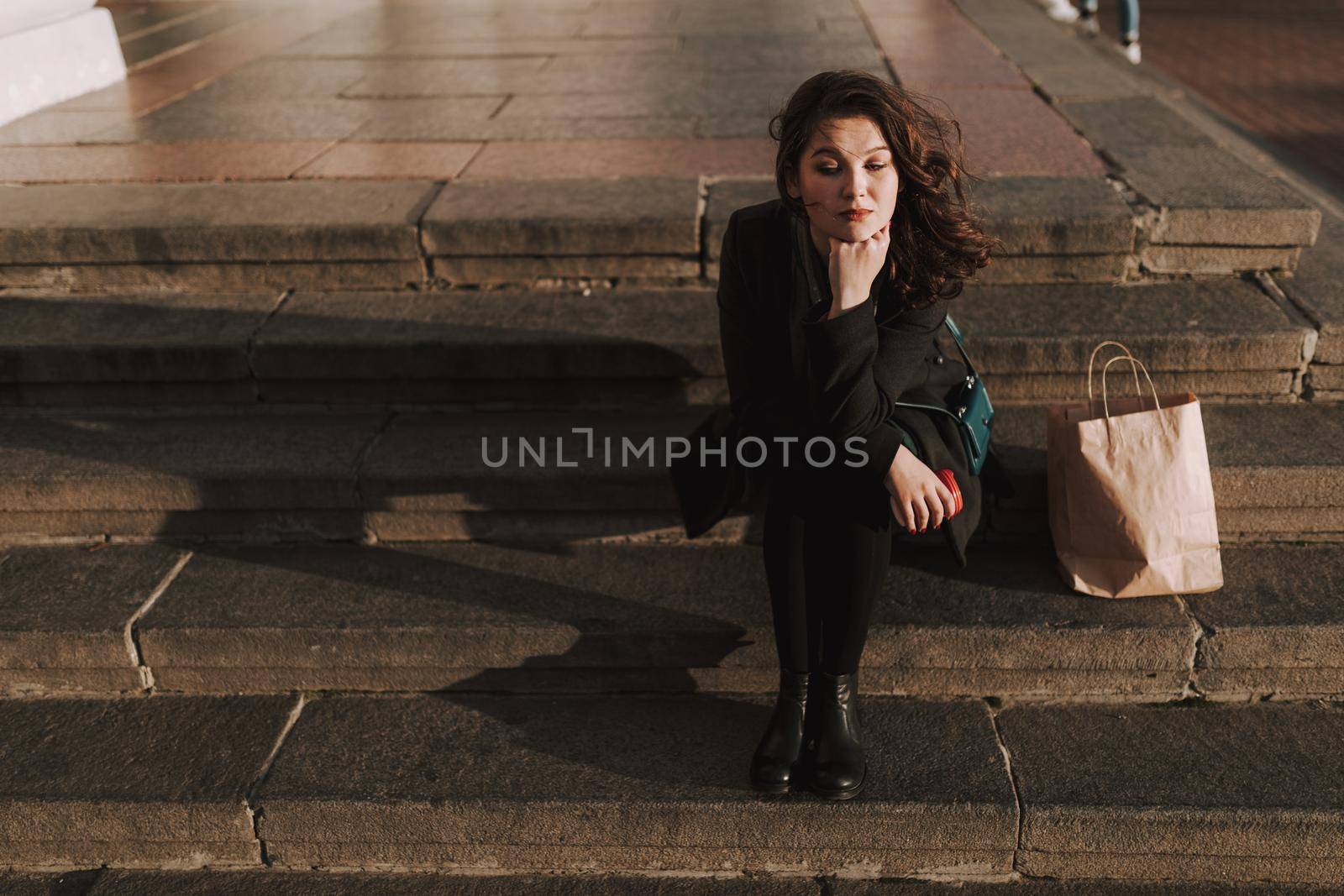 Smiling attractive lady sitting on steps with coffee and shopping bag next to her outdoors