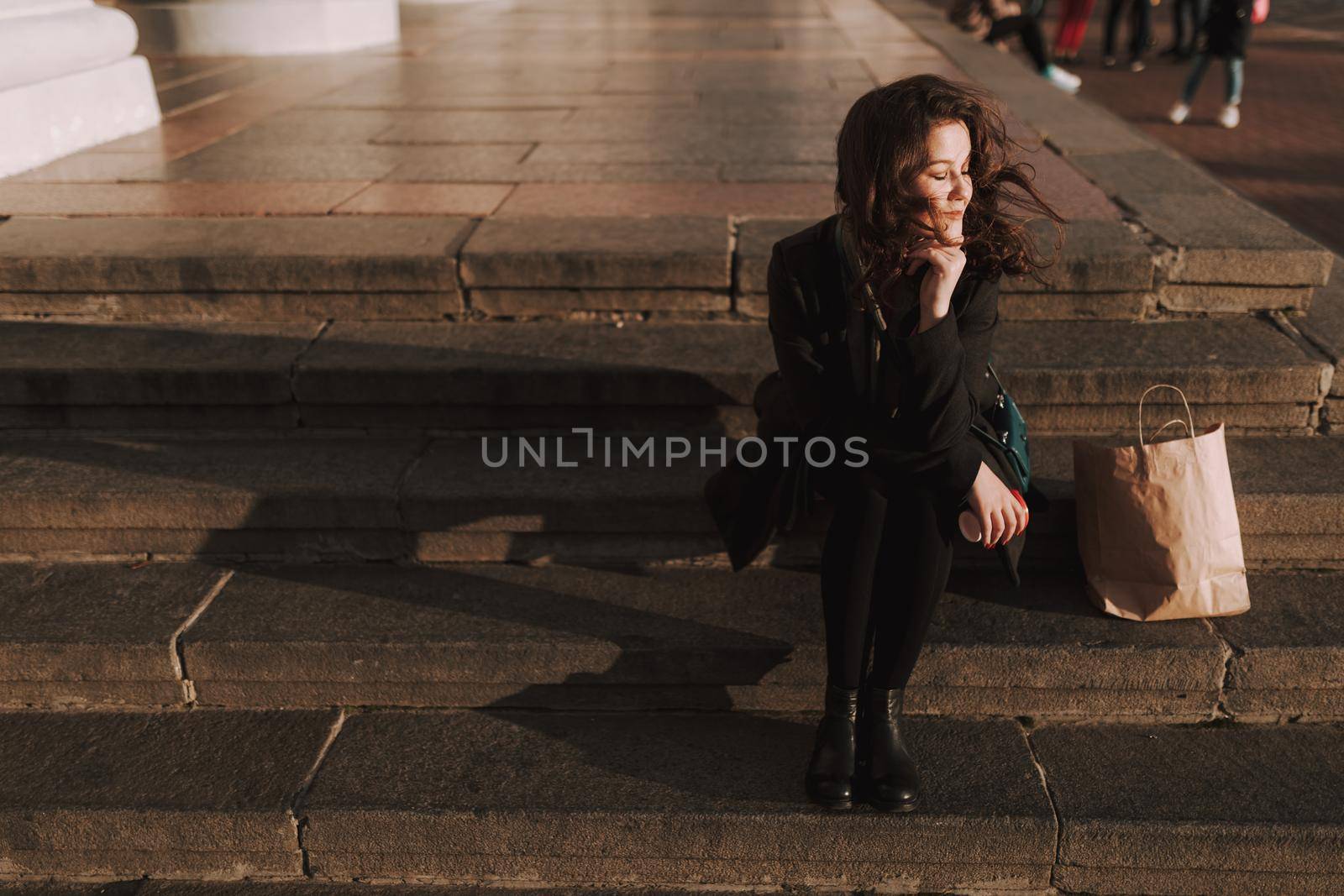 Pretty woman sitting on steps with hot drink and shopping bag next to her outdoors