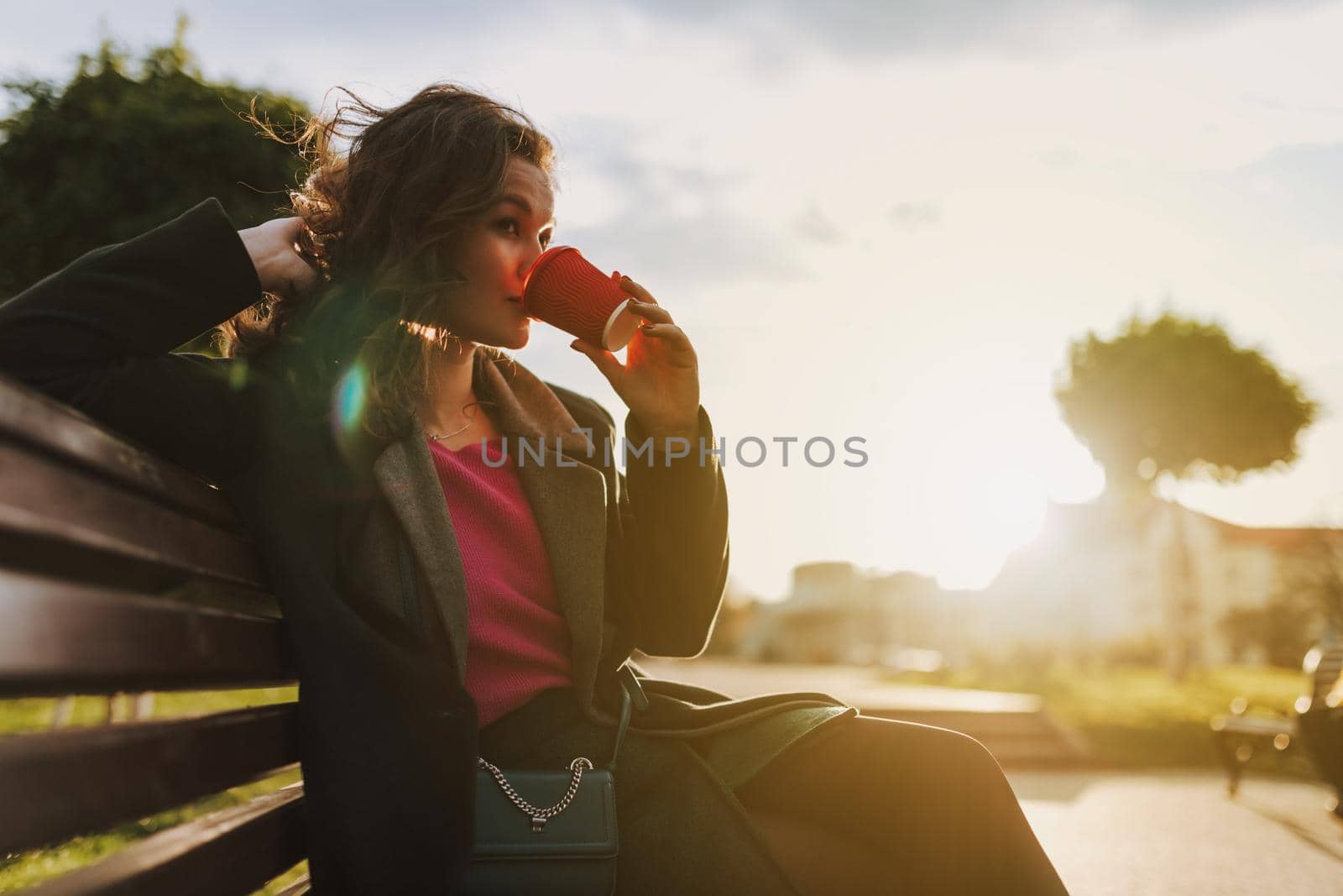 Happy attractive female sitting on bench while holding paper cup of hot drink outdoors