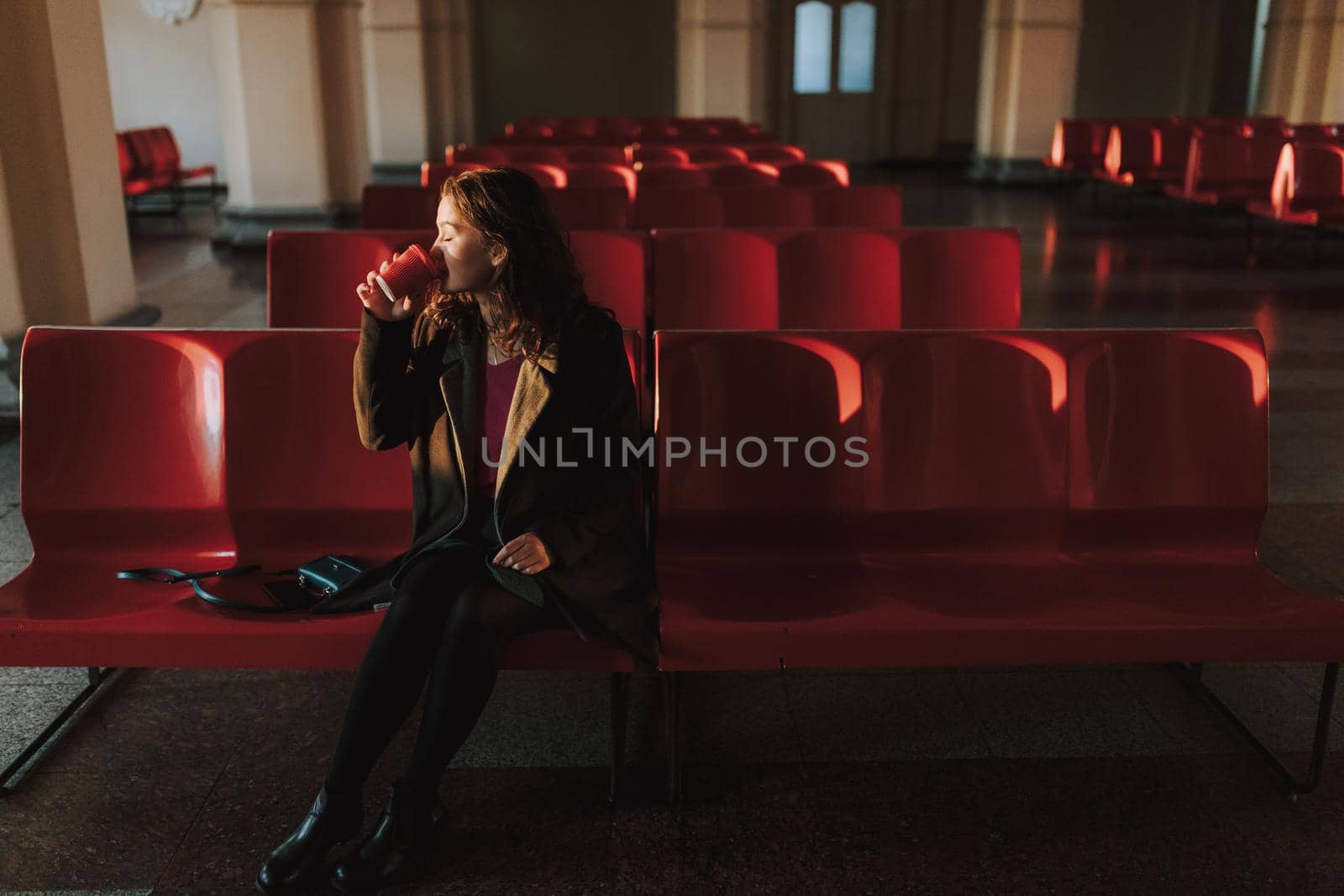 Pretty woman in coat sitting in waiting room on station while holding cup of hot drink