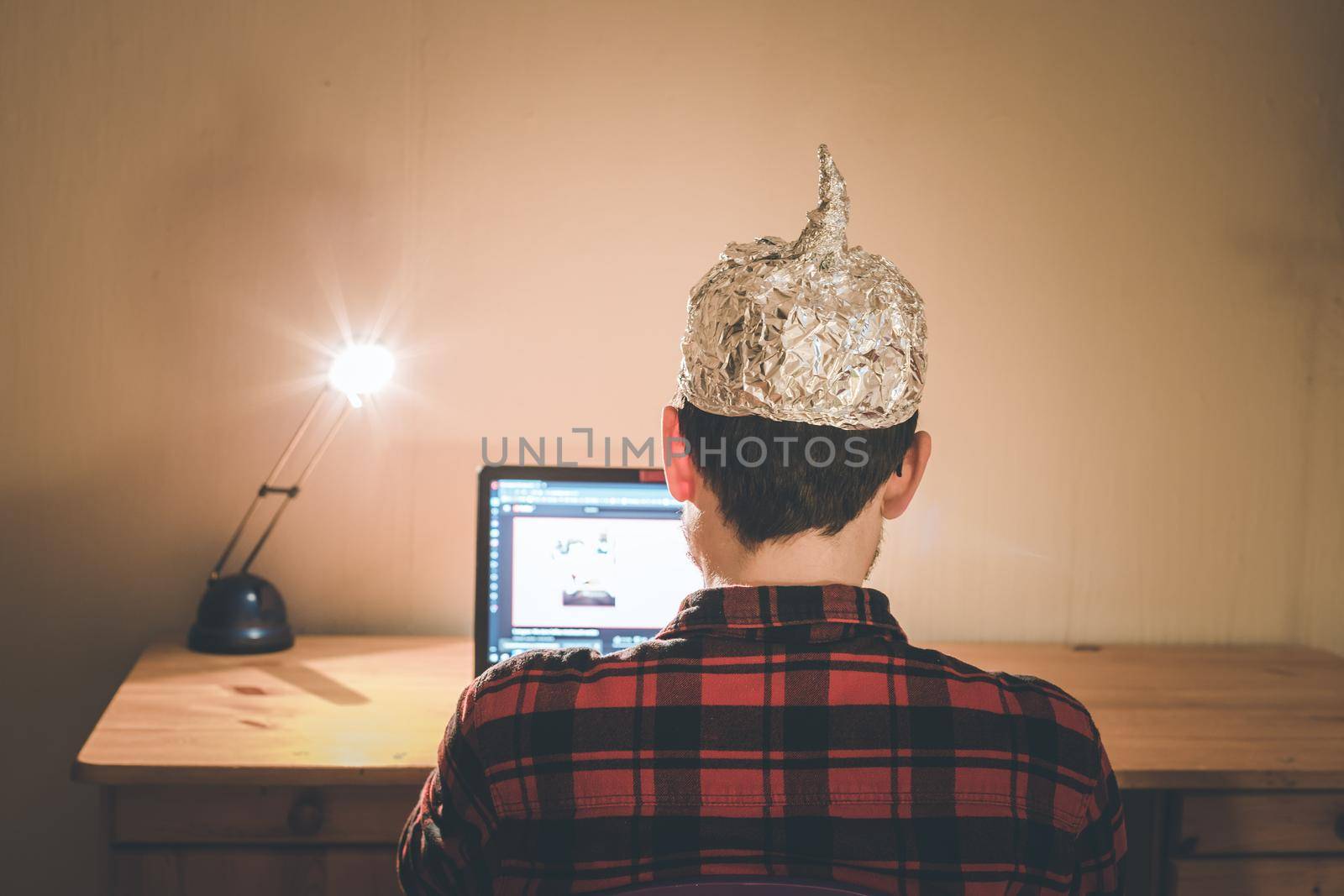 Young man with aluminum cap is sitting in the dark basement in front of a laptop. Conspiracy theory concept