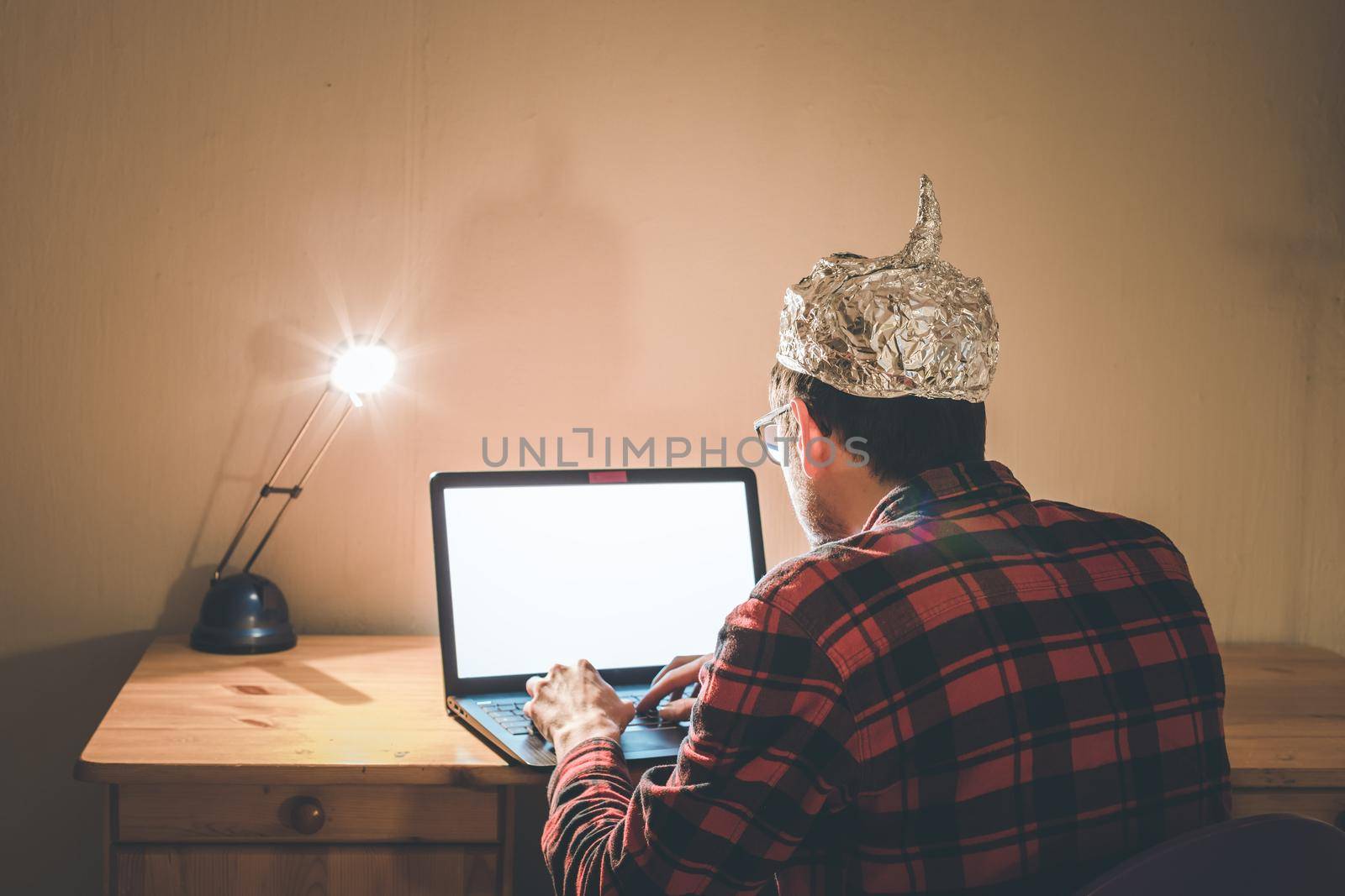 Young man with aluminum cap is sitting in the dark basement in front of a laptop. Conspiracy theory concept