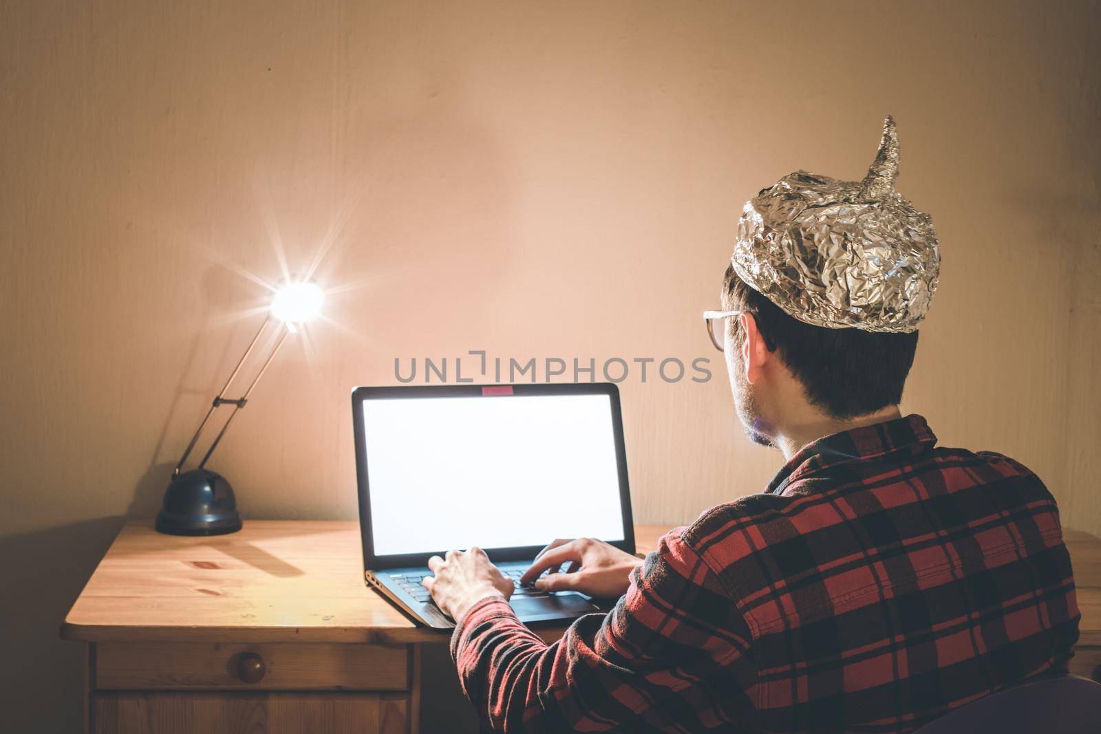 Young man with aluminum cap is sitting in the dark basement in front of a laptop. Conspiracy theory concept
