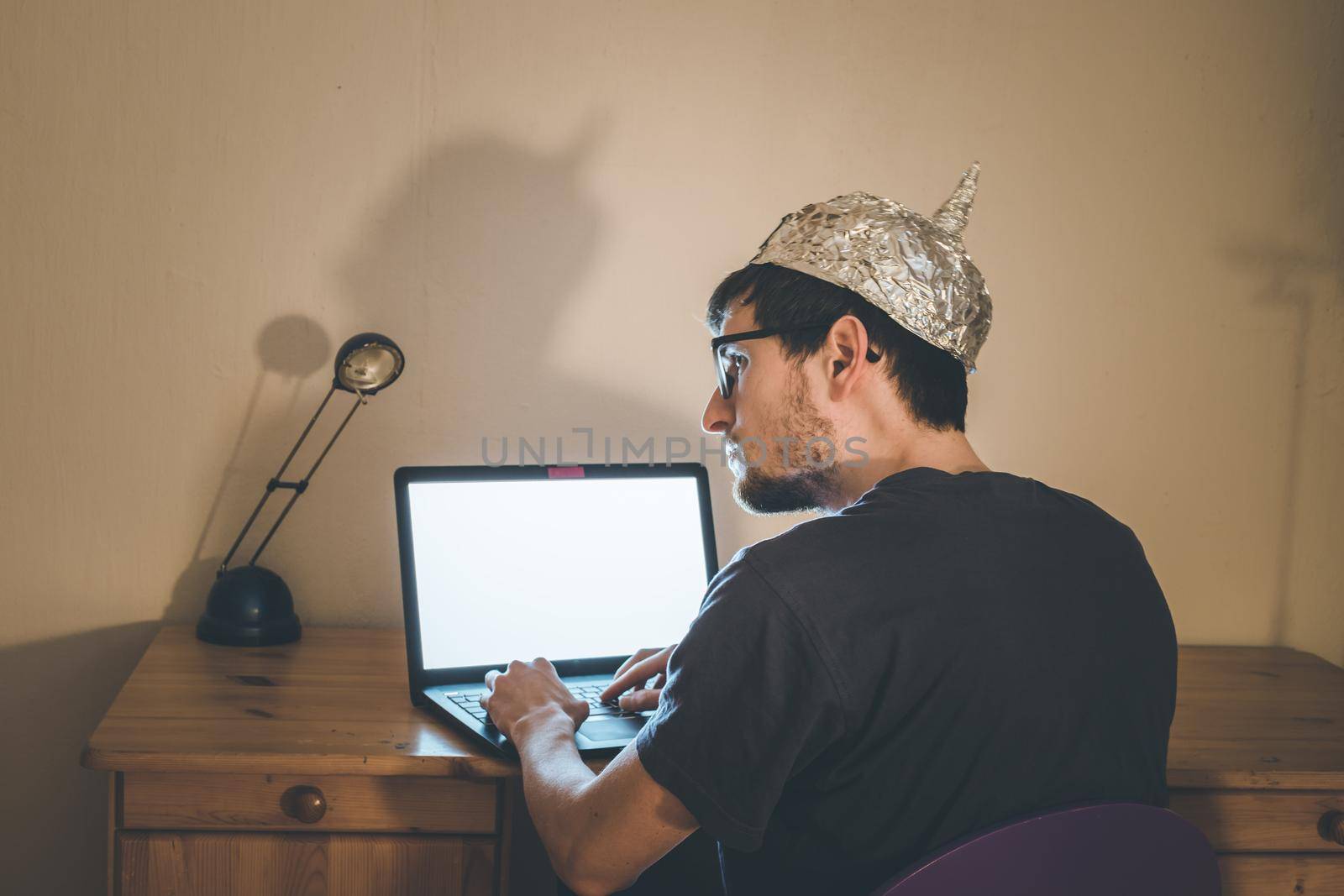 Young man with aluminum cap is sitting in the dark basement in front of a laptop. Conspiracy theory concept