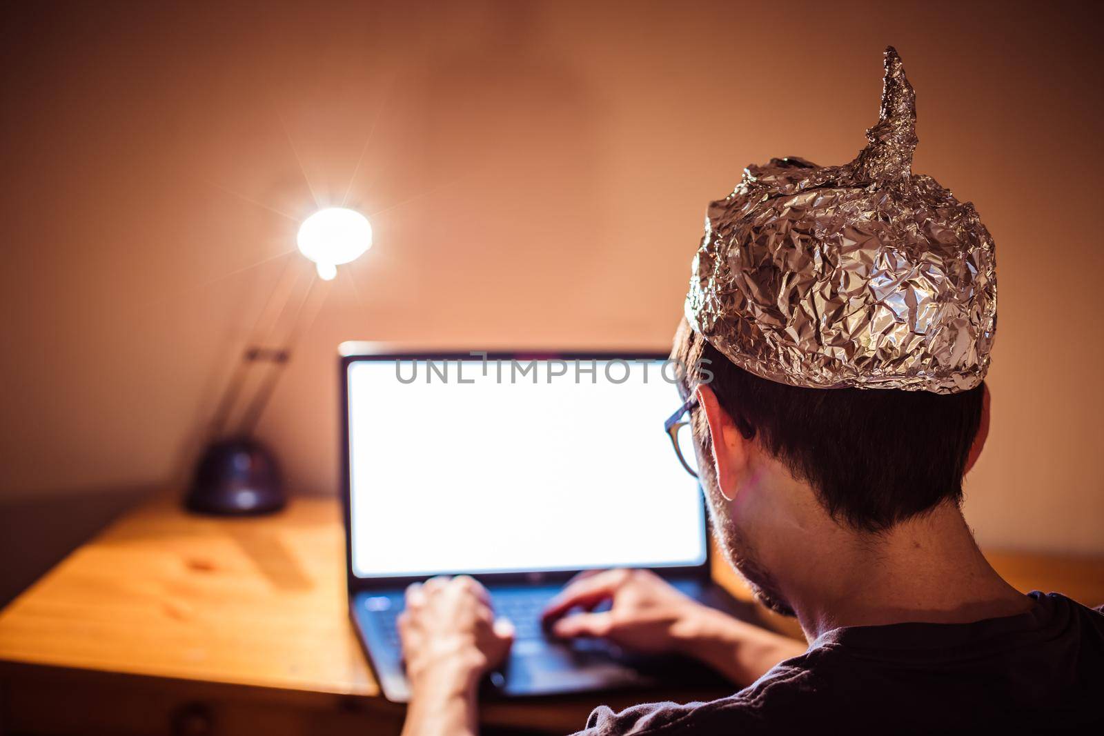 Young man with aluminum cap is sitting in the dark basement in front of a laptop. Conspiracy theory concept