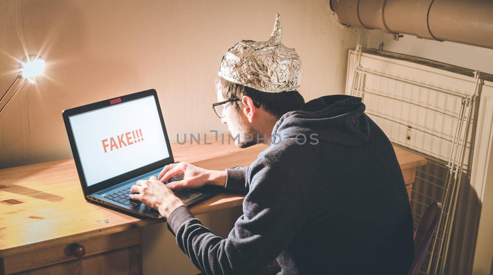 Young man with aluminum cap is sitting in the dark basement in front of a laptop. Conspiracy theory concept