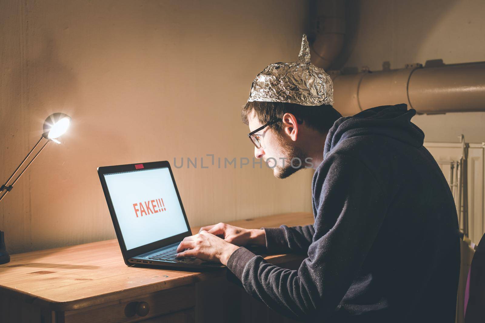 Young man with aluminum cap is sitting in the dark basement in front of a laptop. Conspiracy theory concept