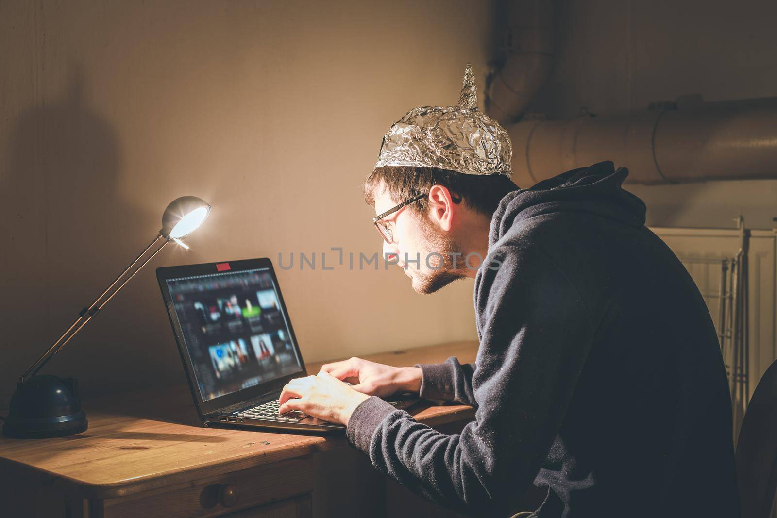 Young man with aluminum cap is sitting in the dark basement in front of a laptop. Conspiracy theory concept