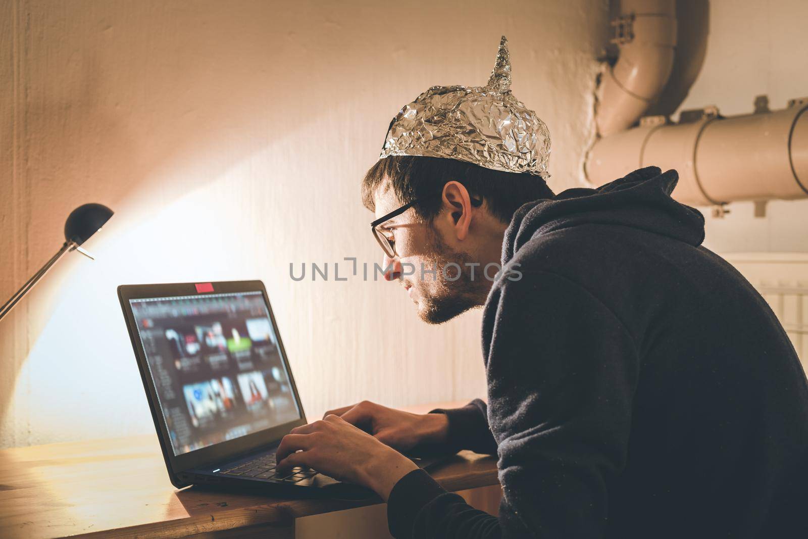 Conspiracy theory concept: young man with aluminum cap searching the internet, sitting lonely in the dark basement by Daxenbichler