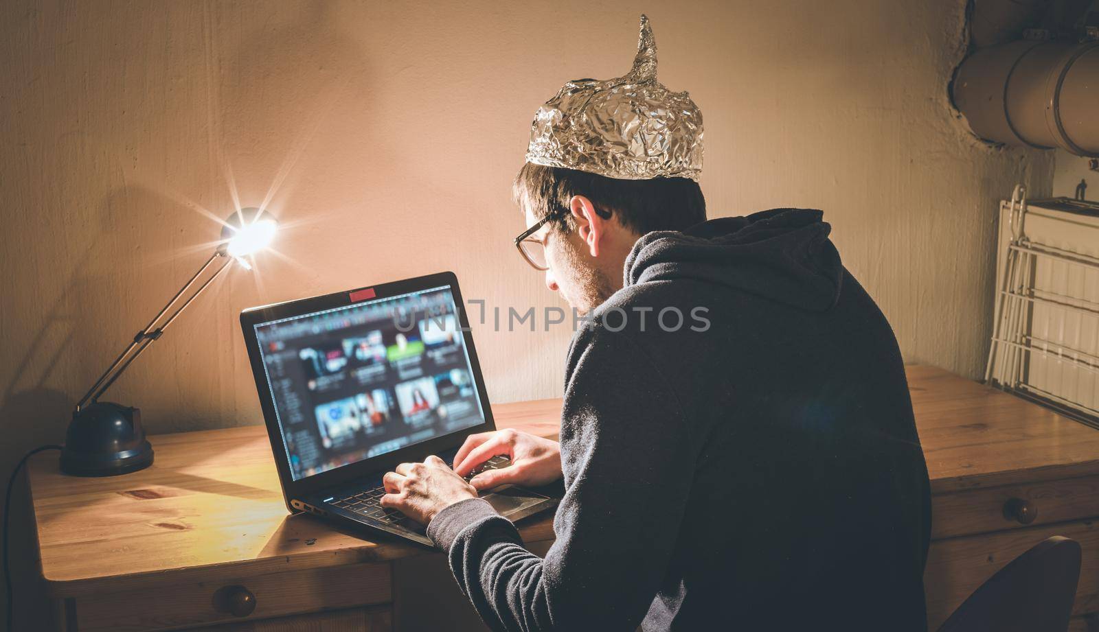 Young man with aluminum cap is sitting in the dark basement in front of a laptop. Conspiracy theory concept