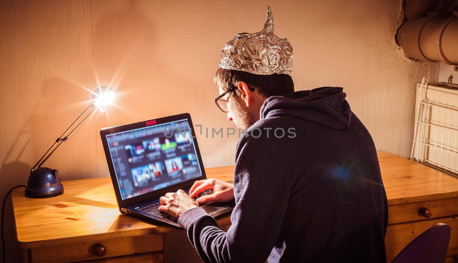 Conspiracy theory concept: young man with aluminum cap searching the internet, sitting lonely in the dark basement by Daxenbichler