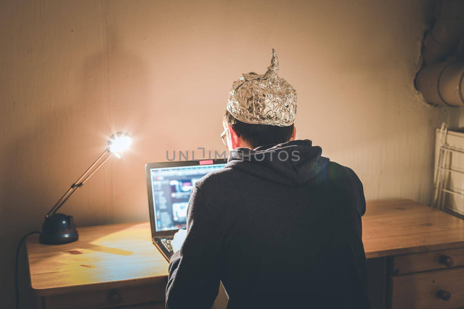 Young man with aluminum cap is sitting in the dark basement in front of a laptop. Conspiracy theory concept