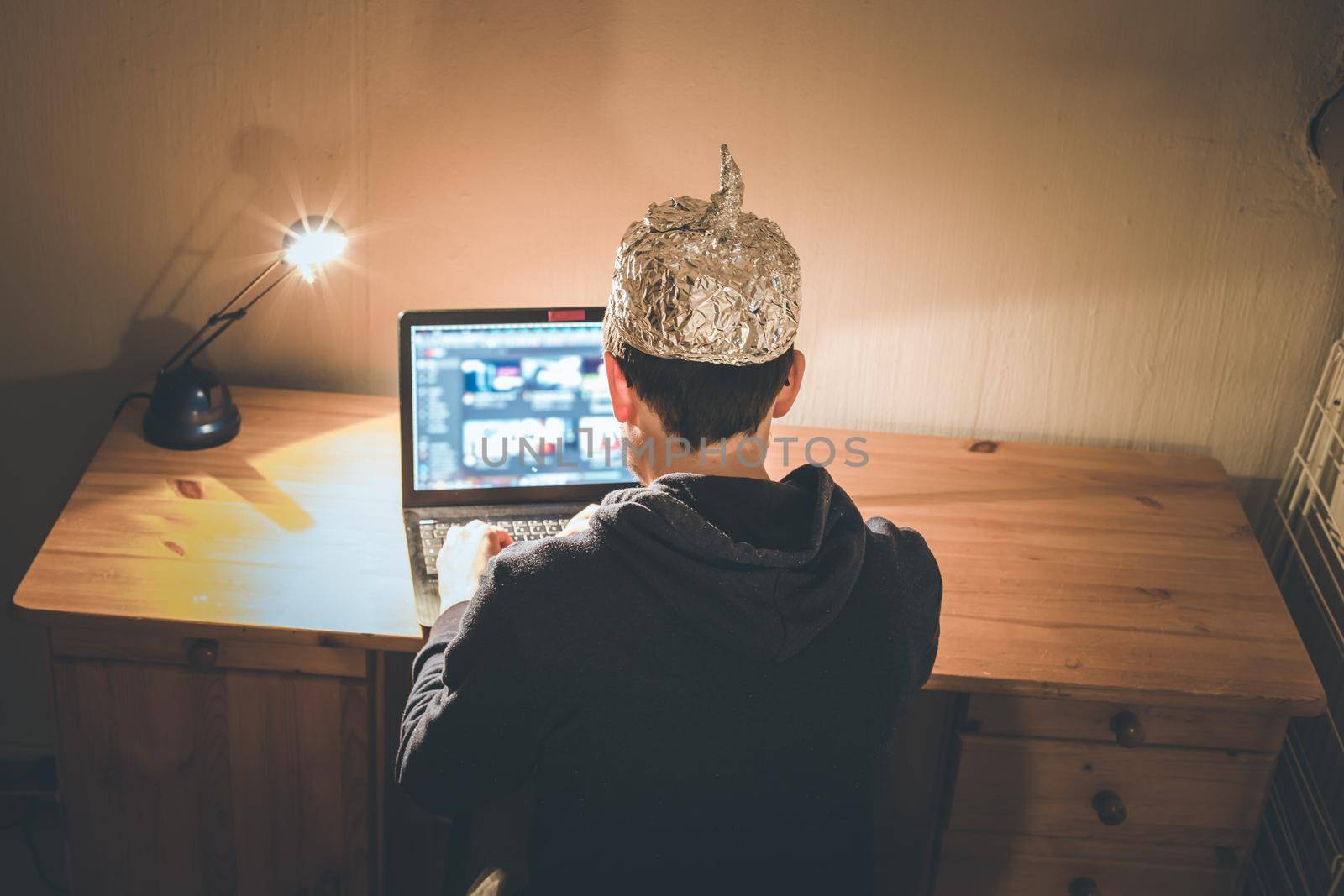 Young man with aluminum cap is sitting in the dark basement in front of a laptop. Conspiracy theory concept