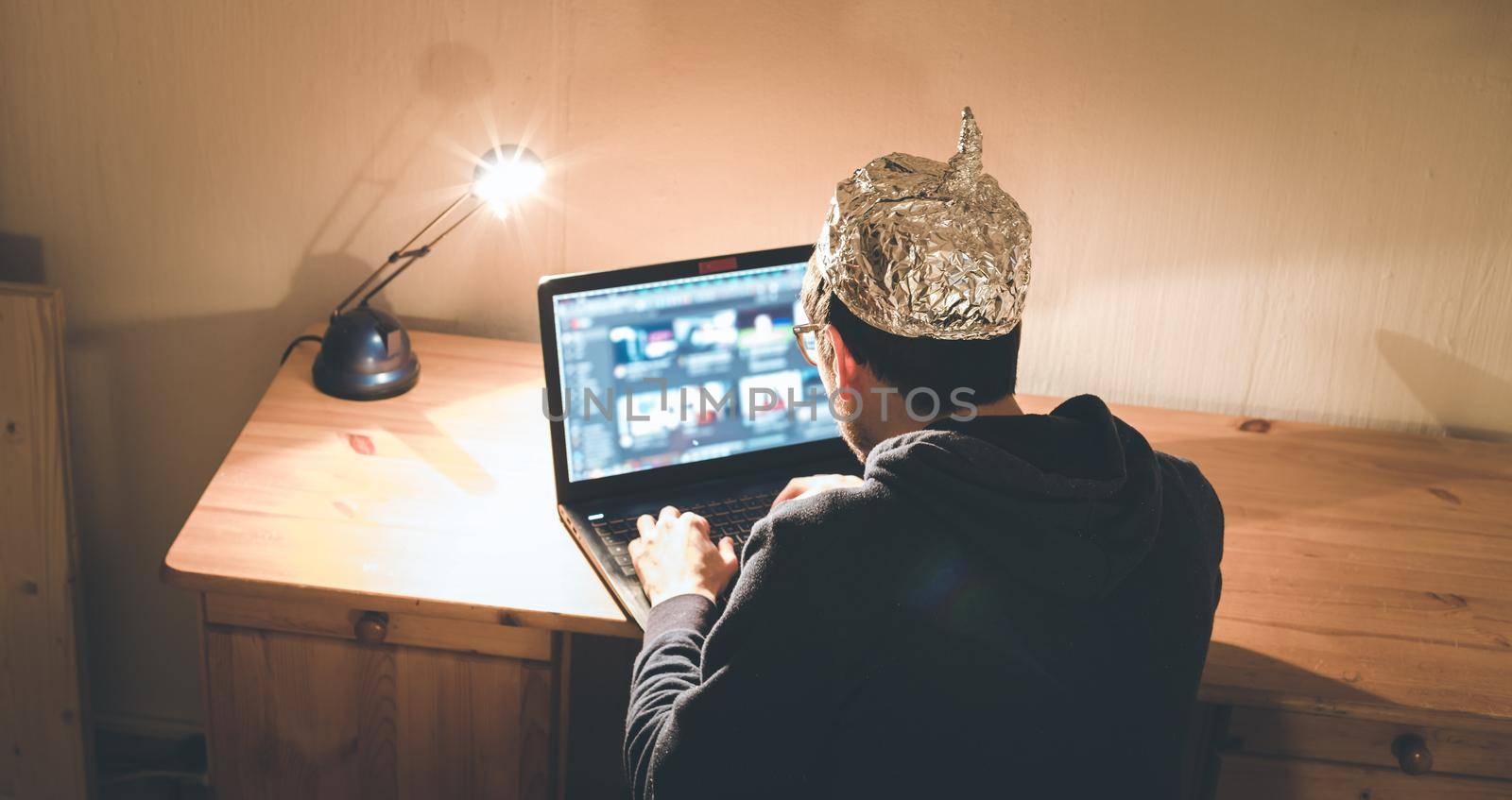 Young man with aluminum cap is sitting in the dark basement in front of a laptop. Conspiracy theory concept