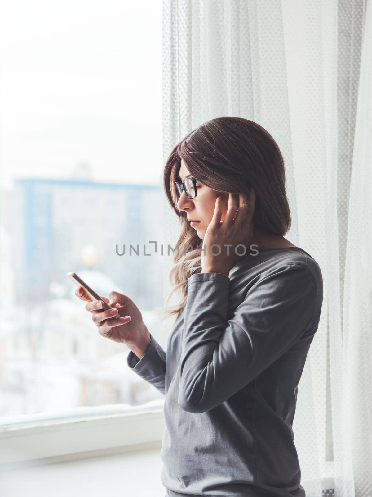 Thoughtful woman with eyeglasses looks at her smartphone. Information in online media. Wireless headphones under hair.