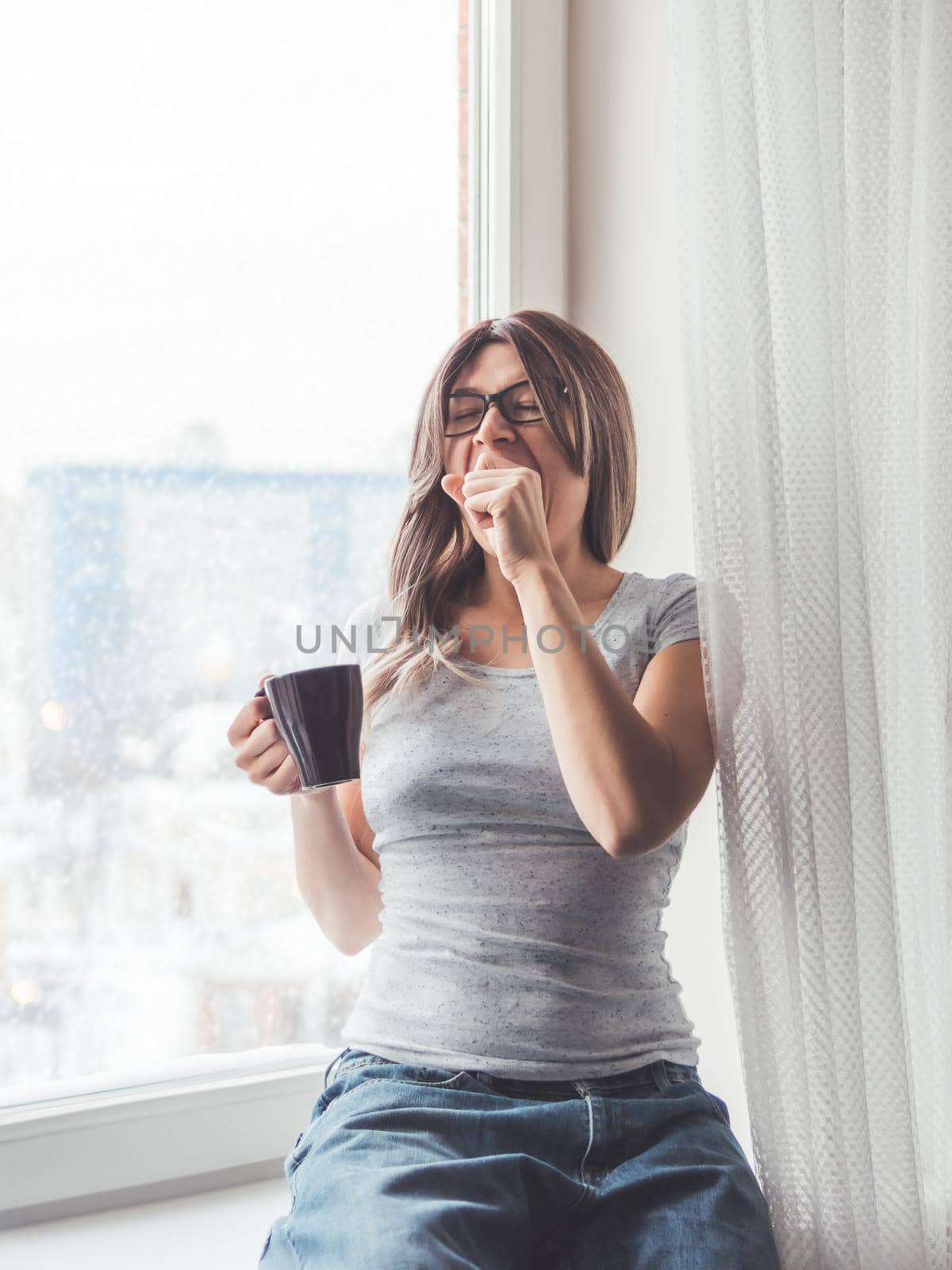 Woman with eyeglasses and curly hair sits on windowsill. Sleepy woman yawns. Morning cup of hot coffee.